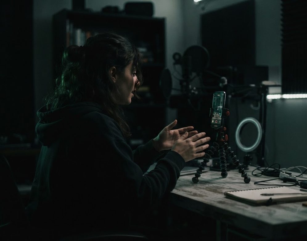 A creator speaking into a studio microphone at a home recording desk, lit by a ring light.